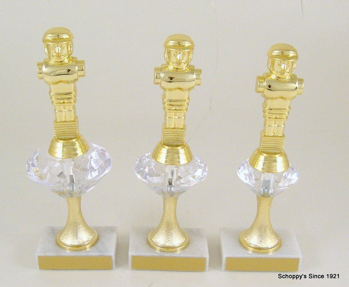 A shiny gold metallic figurine of a foosball figure mounted on a square diamond figure mounted on a white base with a gold front panel, photographed against a plain light background.