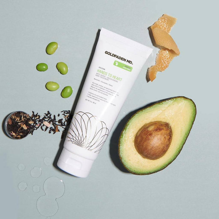 A styled flat lay of Goldfaden MD Hands to Heart cream tube surrounded by its natural ingredients: avocado, edamame, dried tea leaves, and ginger, with water droplets on a sage green surface.