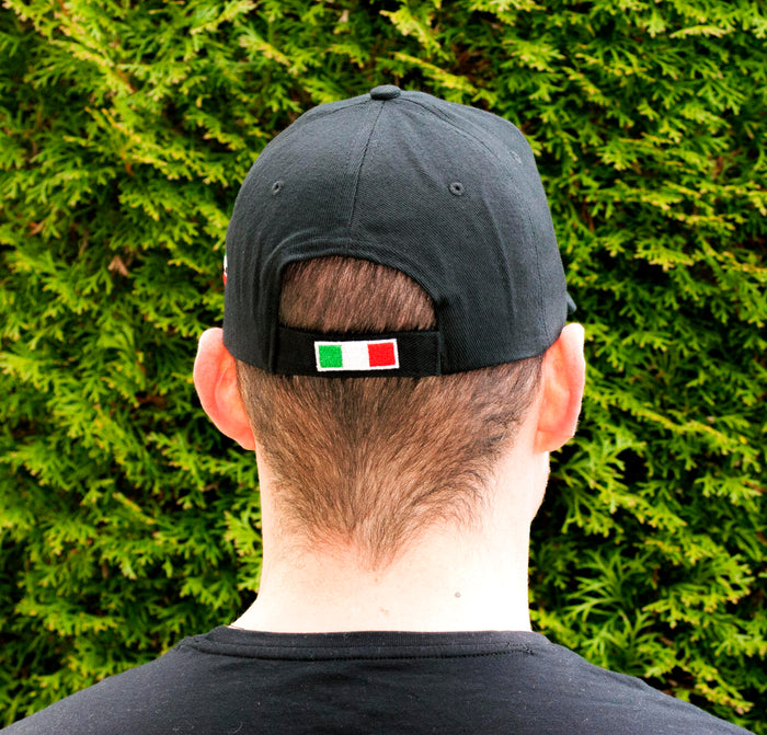 A man wearing a black baseball cap featuring bold, all-caps “ITALIA” embroidery across the front in gray thread, showing the back of the cap. A brushstroke design in green, white, and red—representing the Italian flag—adorns the side, making this cap a stylish tribute to Italian pride and heritage.