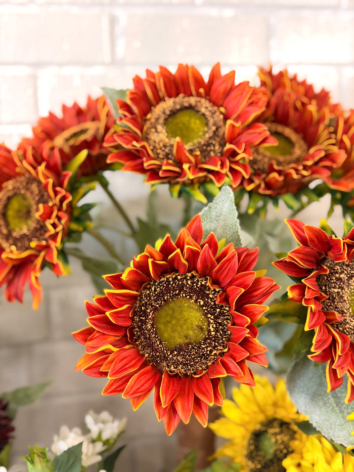 A close-up of artificial sunflower stems with orange-red petals, yellow edging, and dark brown centers, set against a light brick wall background.